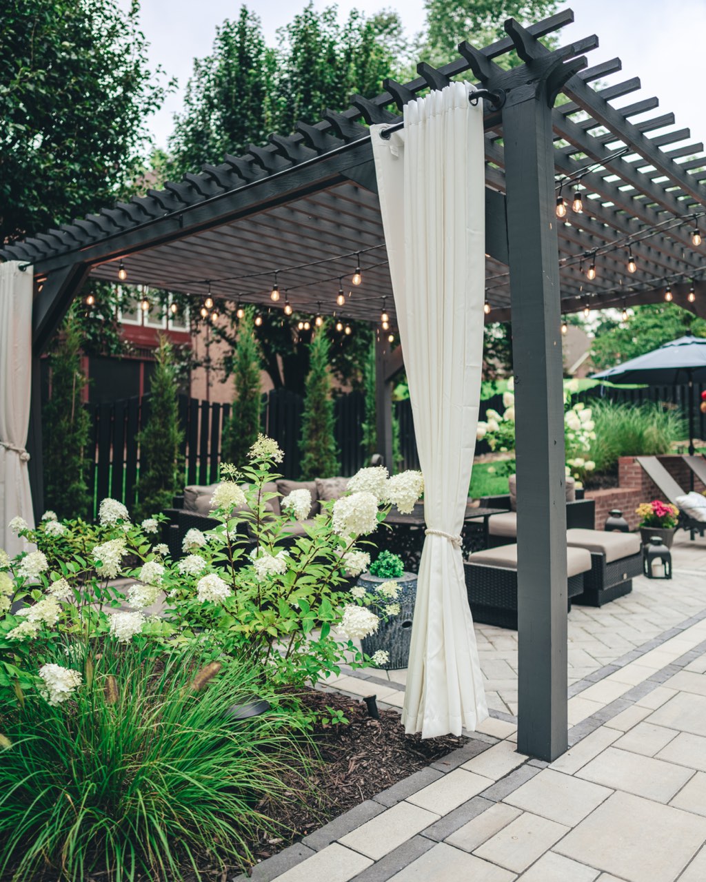 Curtained Pergola Lounge With String Lights And Paver Walkway